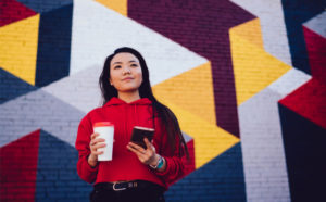 Young Gen Z woman with coffee and phone in front of brightly painted wall