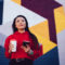 Young Gen Z woman with coffee and phone in front of brightly painted wall